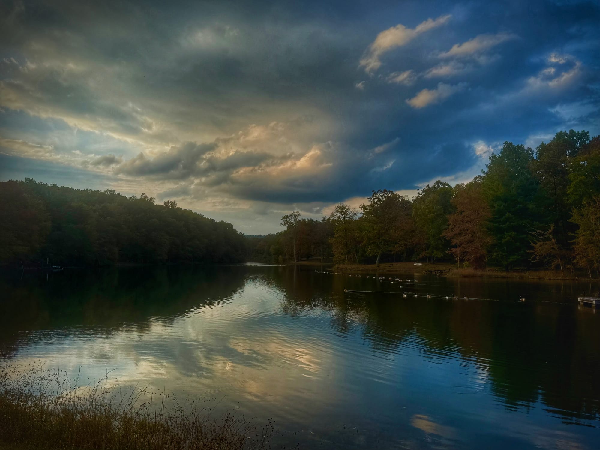 A calm lake reflects towering storm clouds stretching across the evening sky. Dense trees line both shores, their silhouettes framing the water as soft light breaks through gaps in the clouds