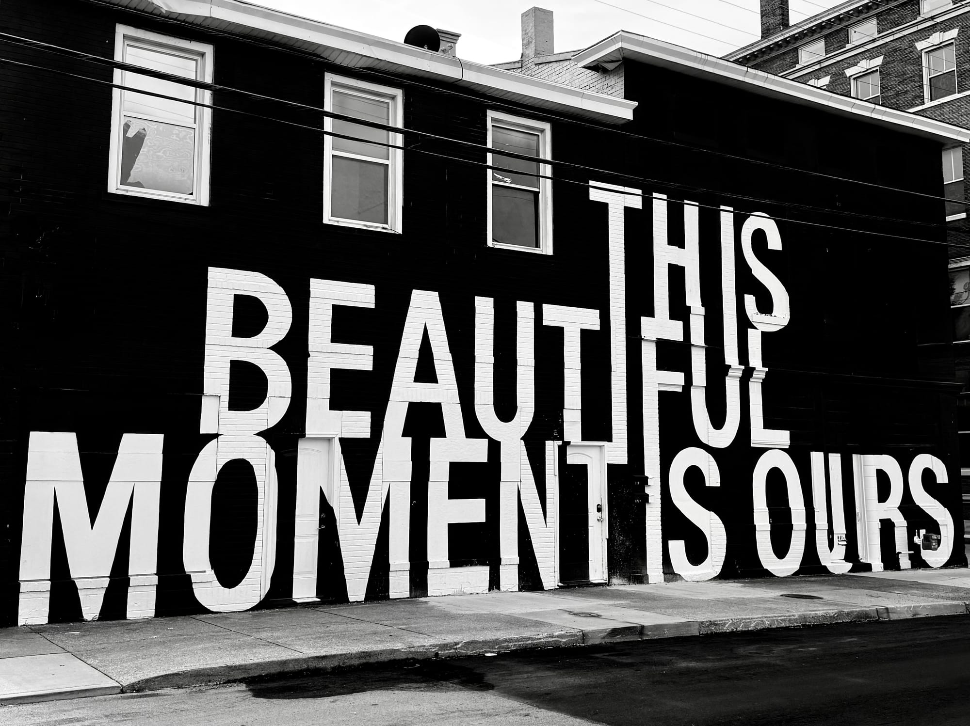 A black and white photo of an urban mural that says in large, bold, white letters "This Beautiful Moment is Ours" on a black wall.